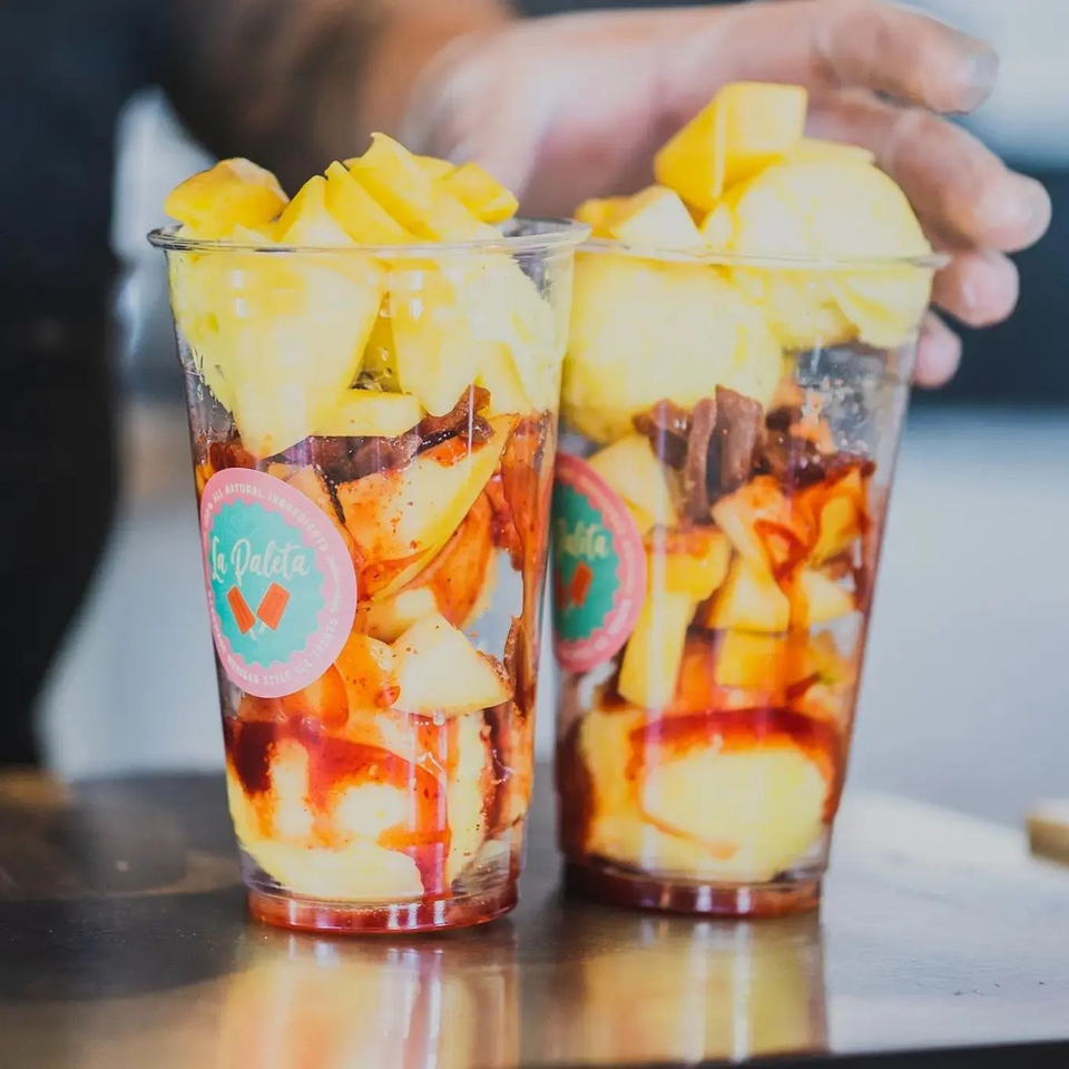 Two La Paleta cups filled with diced mango, chili powder, and chamoy sauce on a counter.