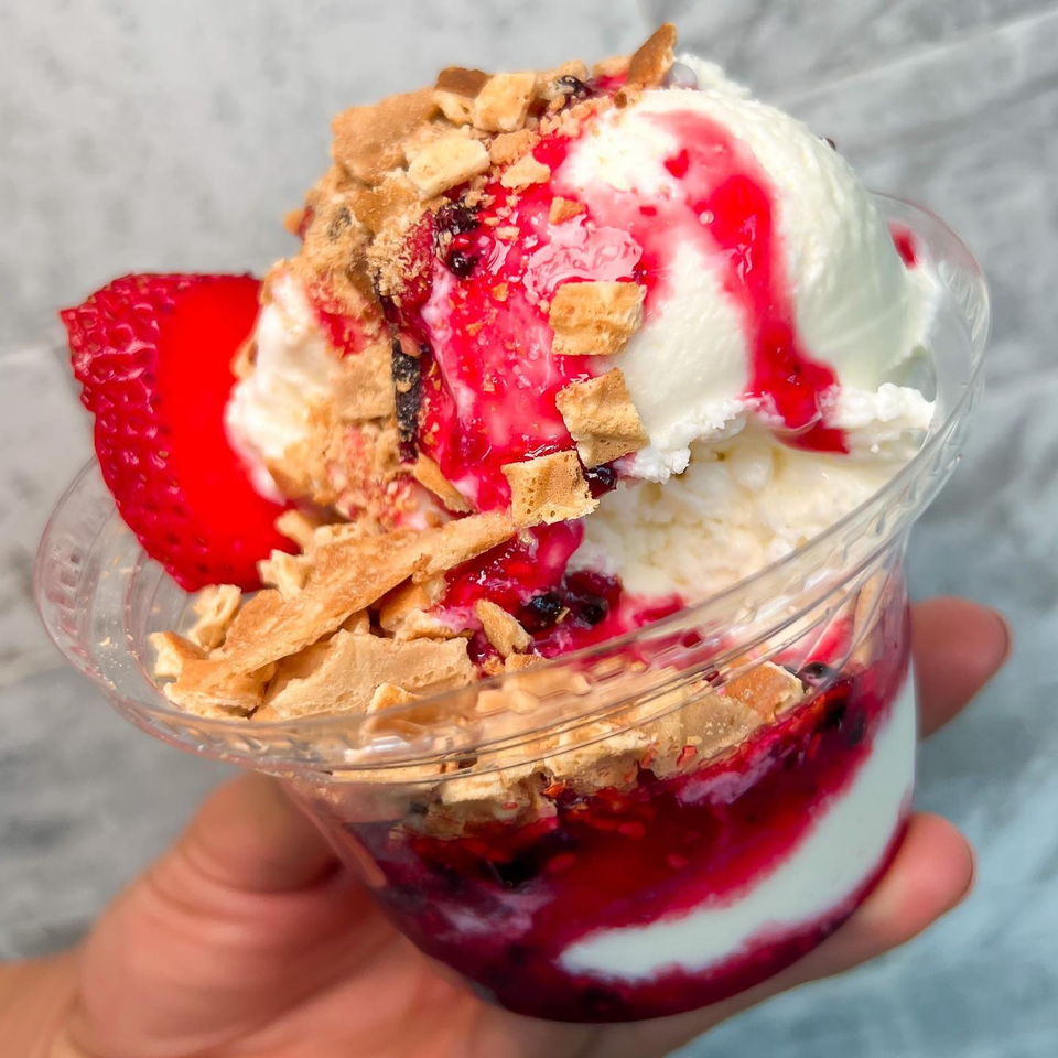 A hand holds a La Paleta dessert cup with vanilla ice cream, berry sauce, and waffle pieces.