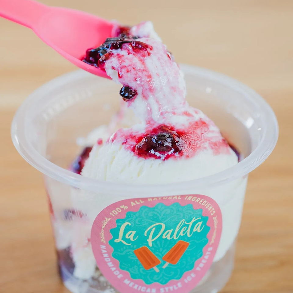 La Paleta ice cream with berry topping in a clear cup, being scooped with a pink spoon.